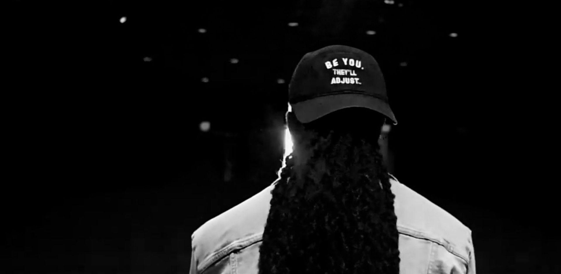 Dr. Okello's back with long hair and ball cap that reads "Be You. They'll Adjust."